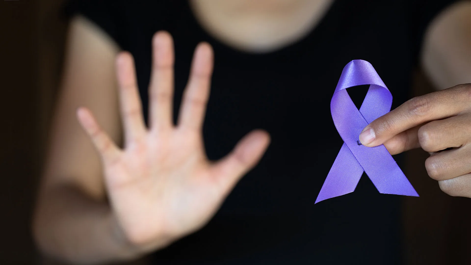 Mujer extendiendo la palma de su mano en señal de alto y con la otra sosteniendo un listón morado, símbolos de erradicar la violencia de género