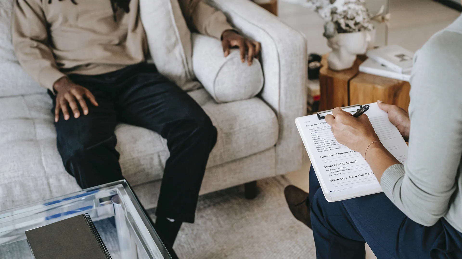 Persona con camisa y pantalón de vestir sentado en un sofá platicando con otra persona enfrente sentada y tomando nota
