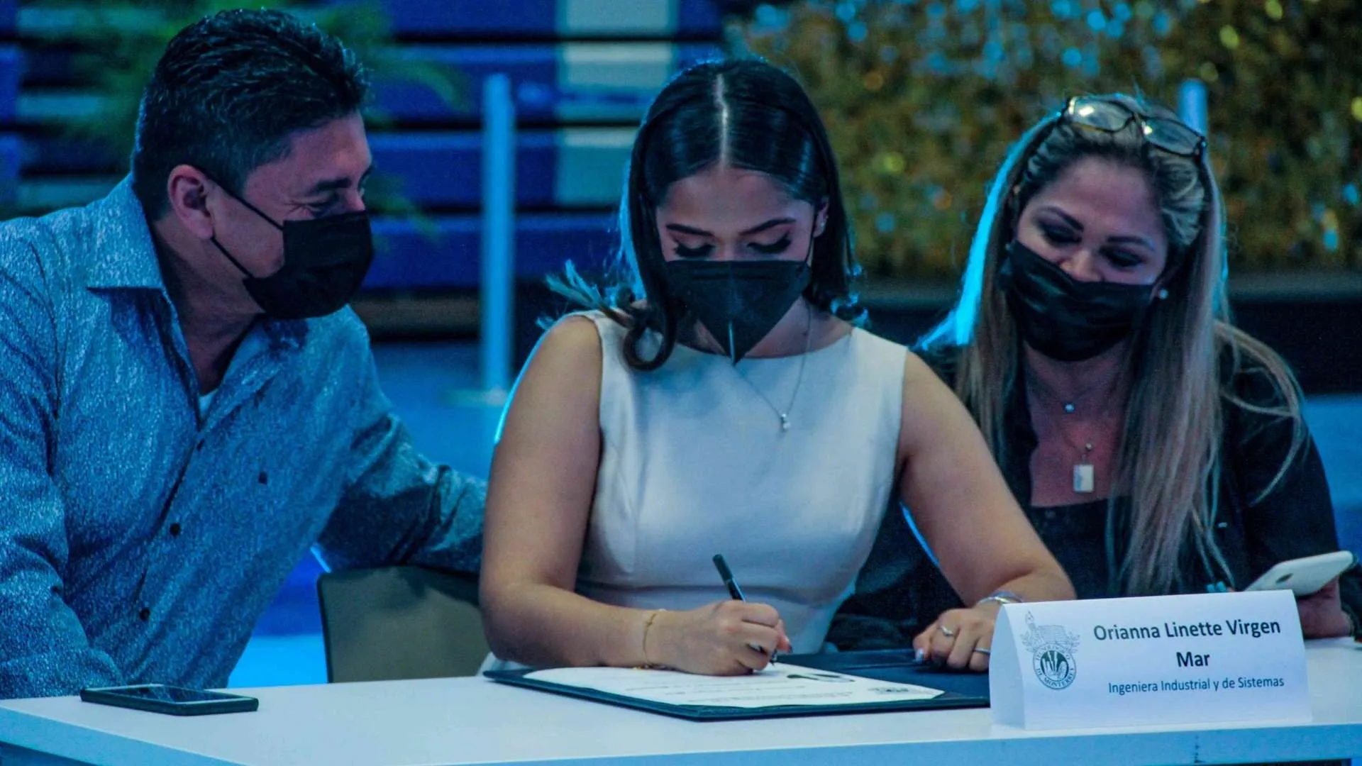 Alumna firmando su título universitario mientras papás observan.