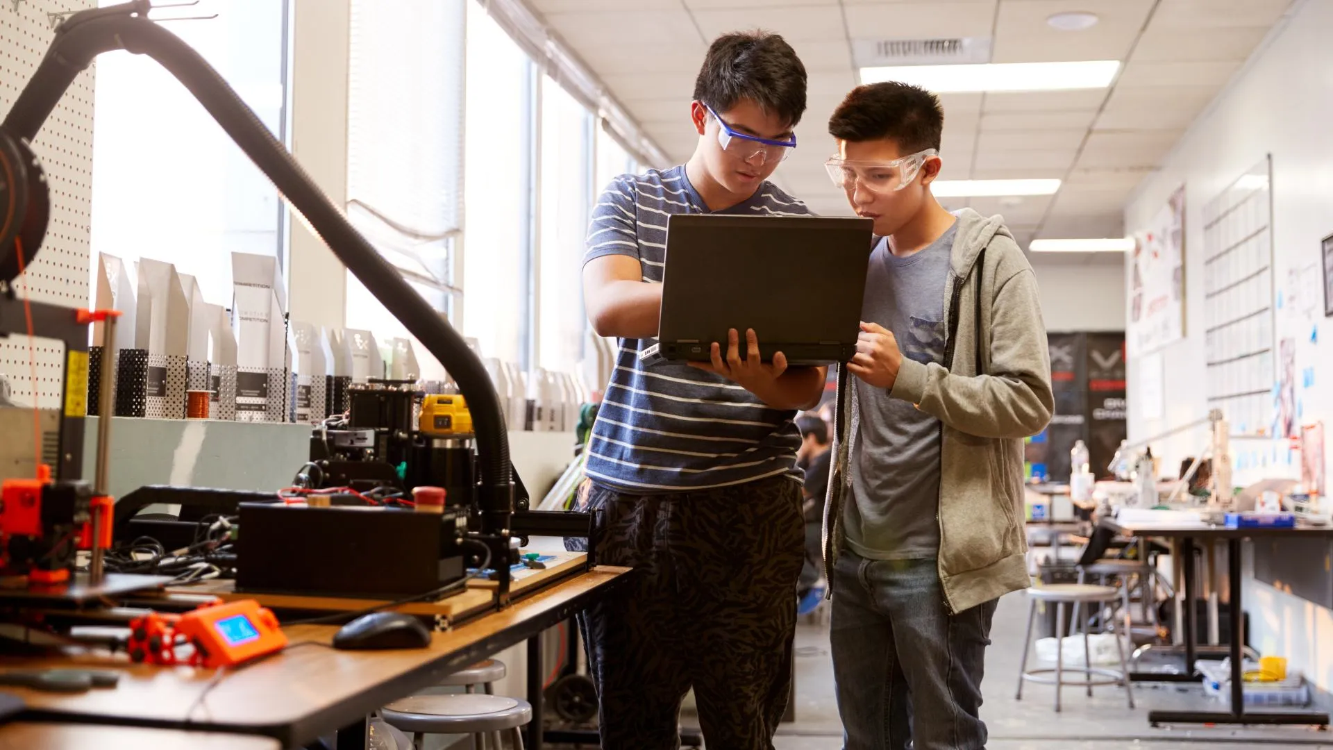 2 jóvenes estudiantes en un laboratorio; uno de ellos sostiene una laptop en su mano y señala su pantalla