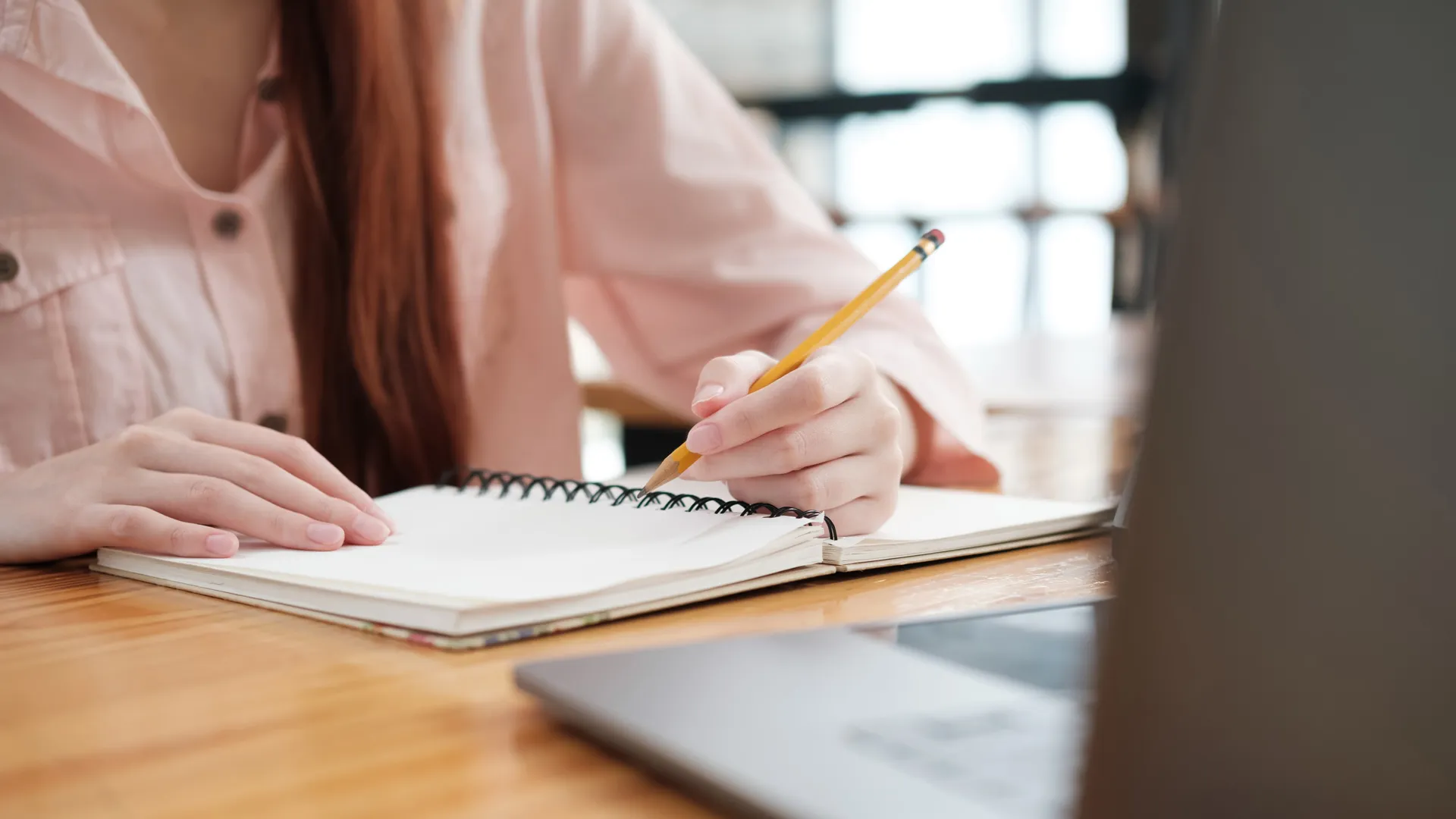 Mujer escribe en su libreta mientras observa su computadora portátil.