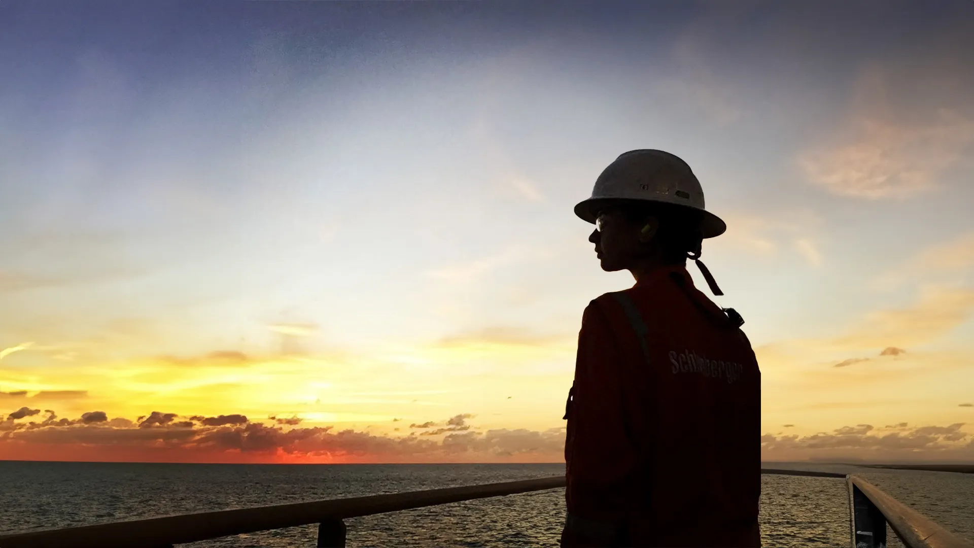 Ashly Hinojos con casco y uniforme naranja mirando el atardecer sobre el mar desde una plataforma petrolera.