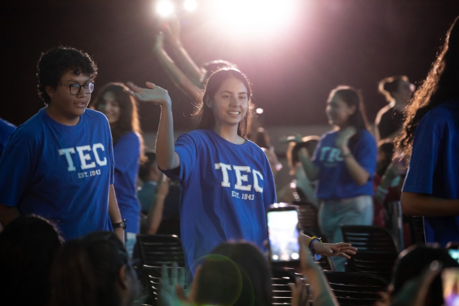 Estudiante con playera azul del Tec sonr&iacute;e y levanta una mano mientras participa junto a sus compa&ntilde;eros en la ceremonia de bienvenida.
