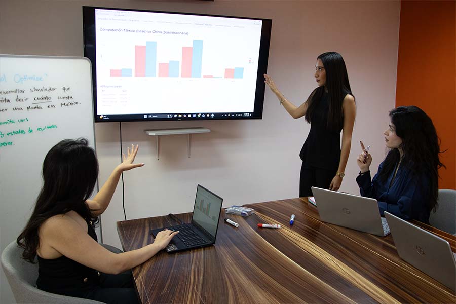 Tres j&oacute;venes en una sala de juntas conversando viendo hacia una pantalla con gr&aacute;ficas de datos. 