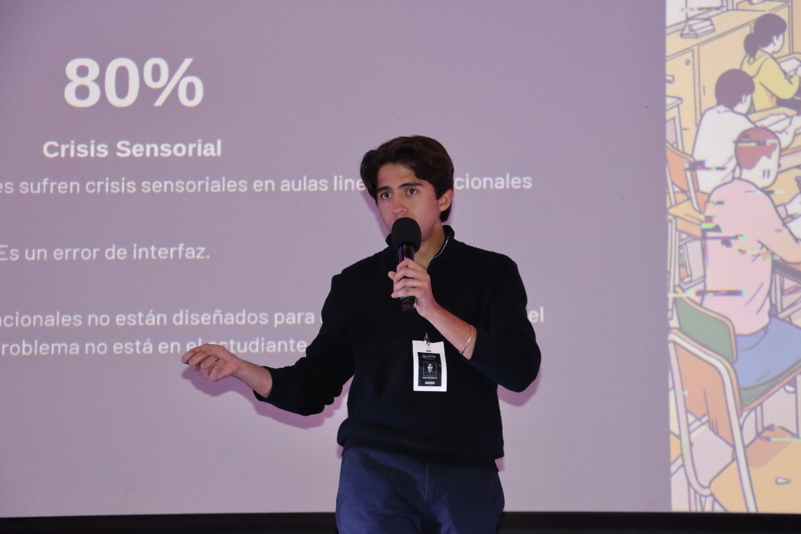Estudiante de PrepaTec Morelia sostiene un micrófono y expone el proyecto Elysium frente a una pantalla que muestra datos sobre crisis sensorial en aulas tradicionales. Detrás se observan gráficos e ilustraciones relacionadas con el entorno escolar, durante su presentación en el Encuentro Nacional de Líderes con Sentido Humano.