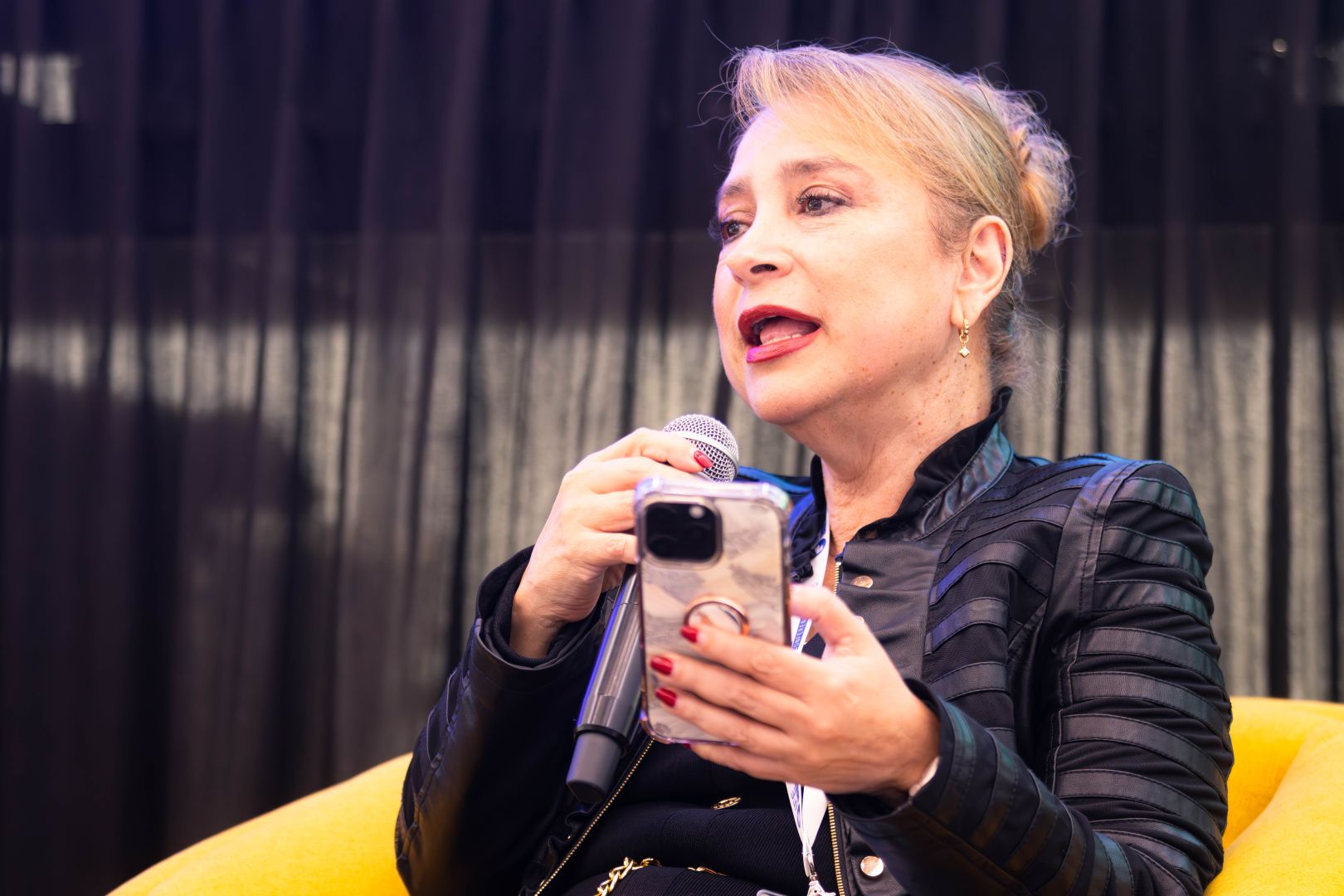 Ponente sentada en un sillón amarillo, con chamarra negra, sostiene un micrófono y un teléfono celular mientras lee y expone sus ideas en un panel educativo.