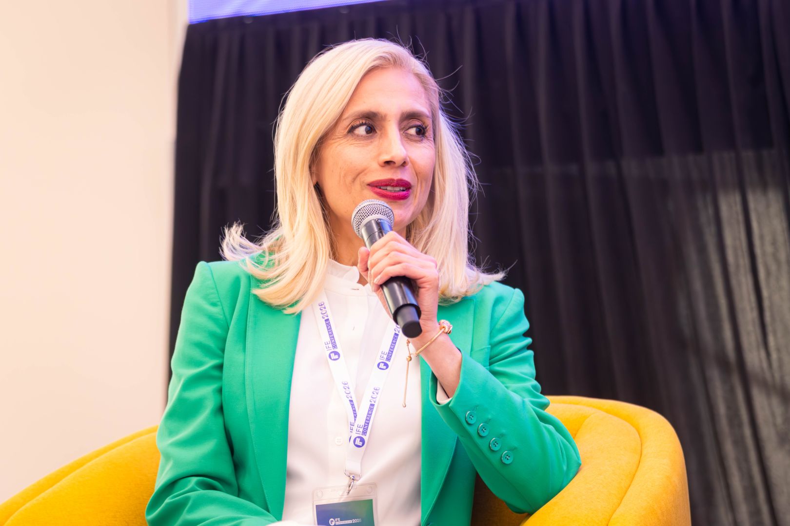 Ponente sentada en un sillón amarillo, vestida con saco verde y blusa blanca, sostiene un micrófono mientras participa en un panel sobre innovación educativa frente a una audiencia.