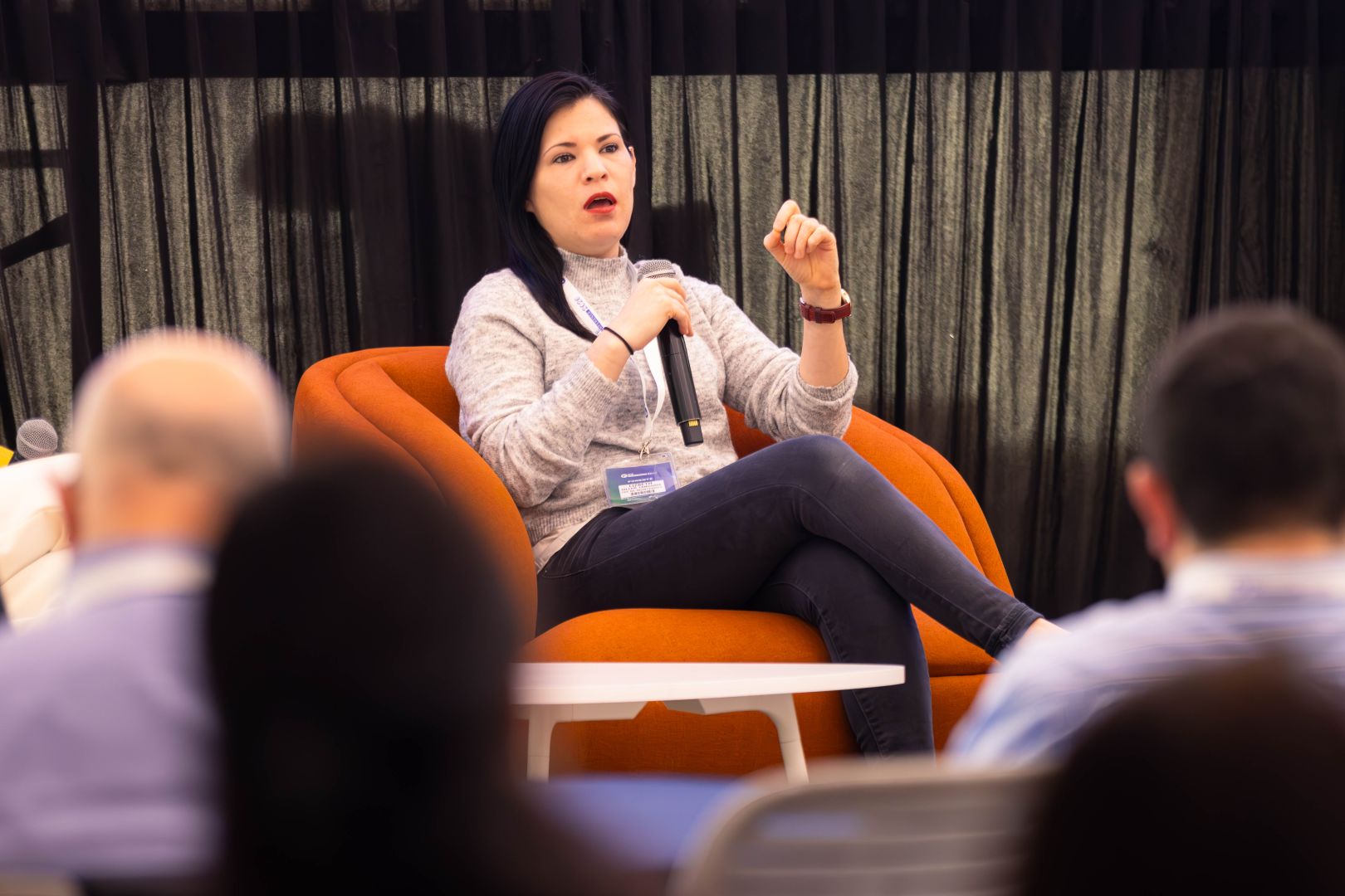 Ponente sentada en un sillón naranja, vestida con suéter gris, sostiene un micrófono y expone sus ideas durante un panel educativo ante una audiencia.