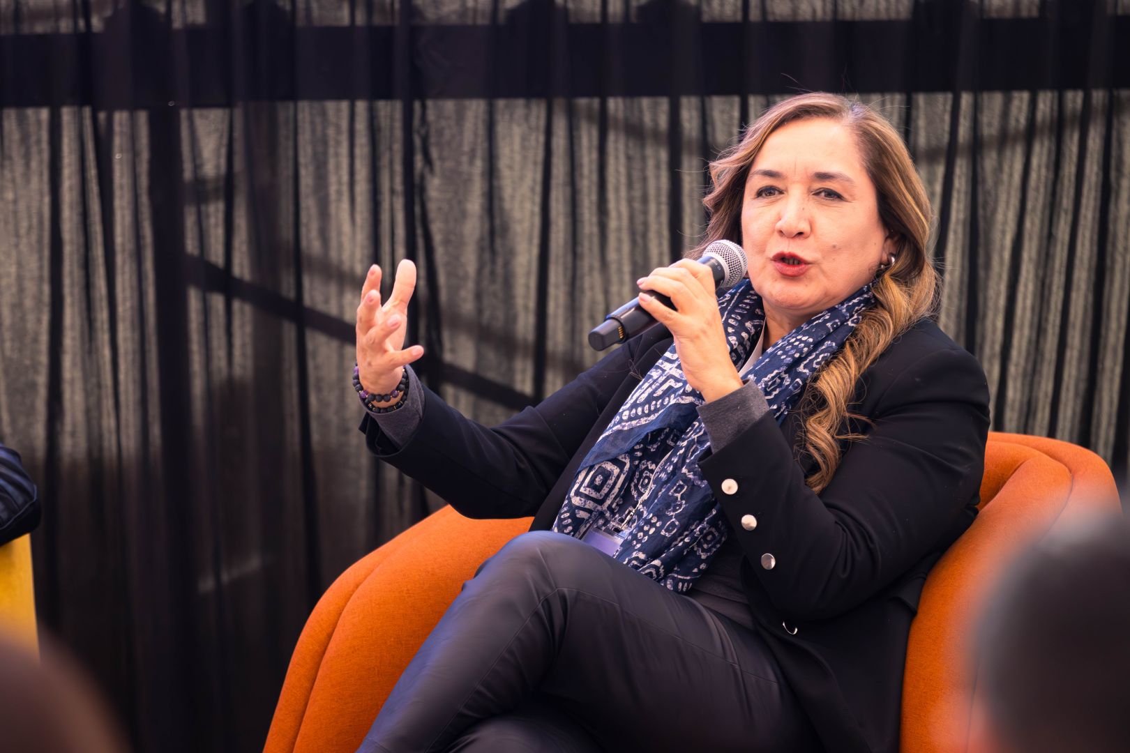 Ponente sentada en un sillón naranja, con saco oscuro y pañuelo, habla al micrófono mientras gesticula durante un panel frente a asistentes.