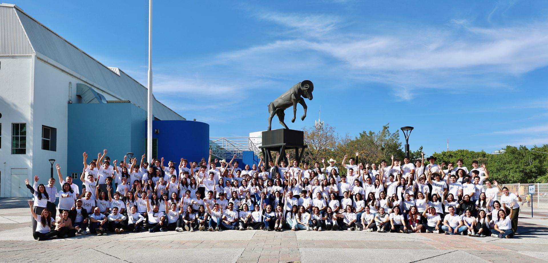 Fotografía panorámica de un gran grupo de estudiantes y profesores de PrepaTec vestidos con playeras blancas, reunidos al aire libre en un campus universitario. Las y los participantes levantan las manos y sonríen frente a una escultura emblemática, reflejando un ambiente de celebración, comunidad y participación colectiva.
