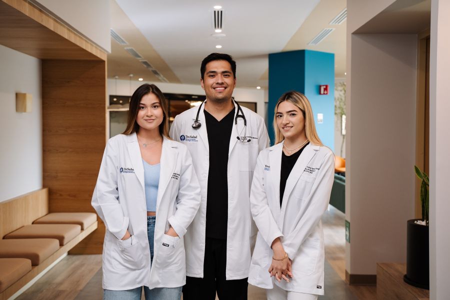 Tres estudiantes de Medicina con bata blanca posan en un espacio cl&iacute;nico-acad&eacute;mico de la Escuela de Medicina y Ciencias de la Salud.