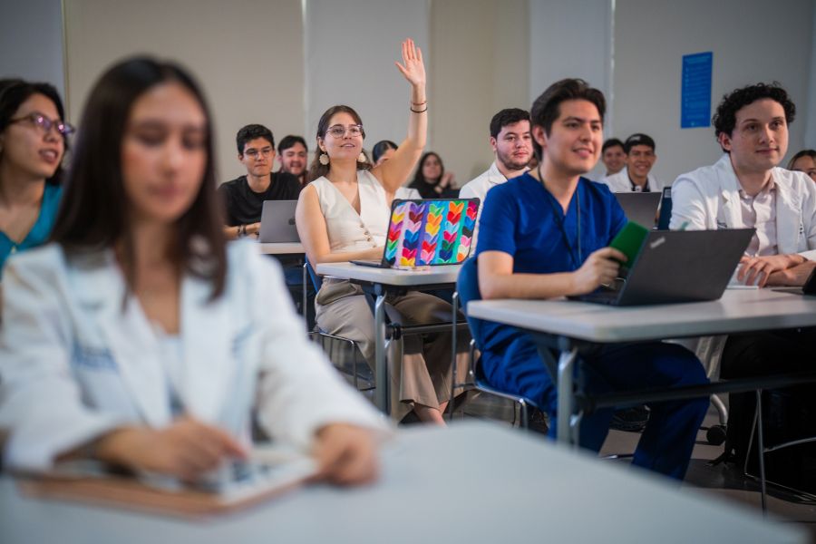 Estudiantes de Medicina en un sal&oacute;n de clases; una alumna levanta la mano para participar mientras compa&ntilde;eros toman apuntes y utilizan computadoras port&aacute;tiles.