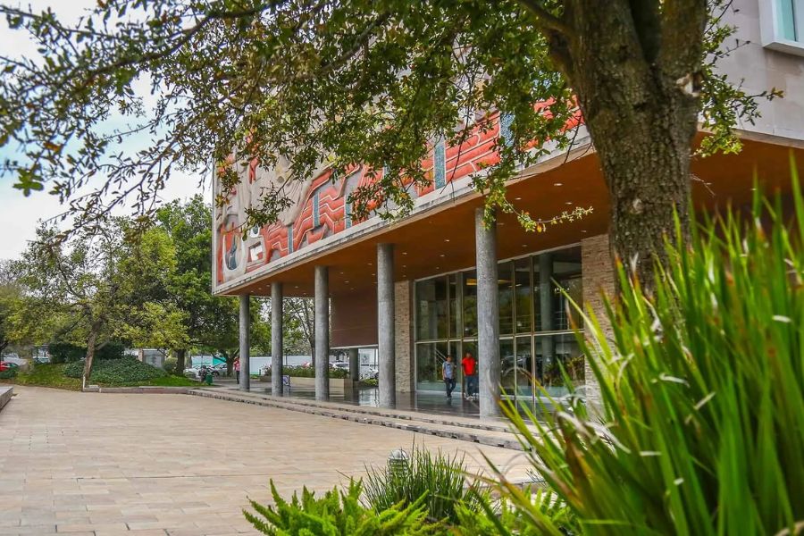 Vista exterior de un edificio del Tecnol&oacute;gico de Monterrey rodeado de &aacute;reas verdes y &aacute;rboles, con personas caminando hacia la entrada principal del campus.