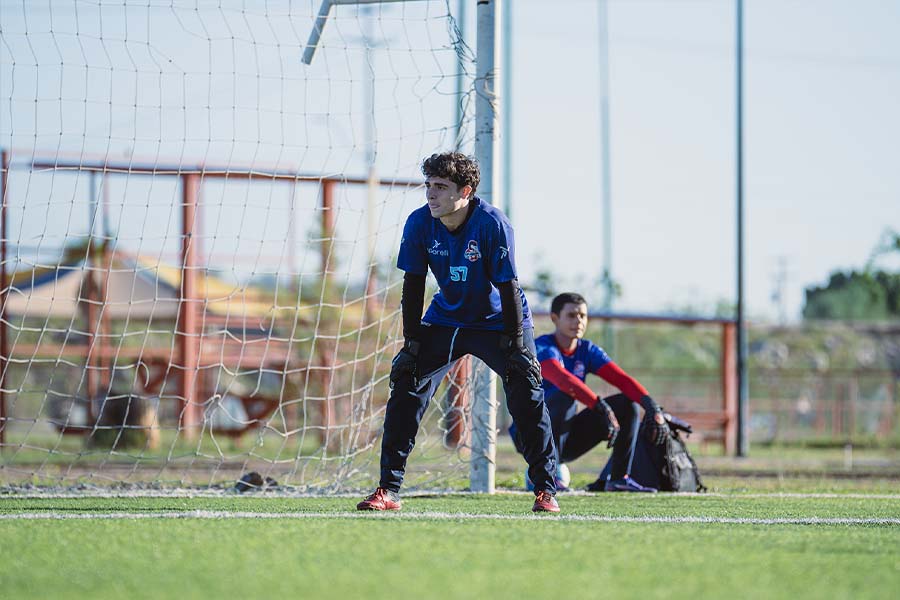 Joven vestido con uniforme de f&uacute;tbol, parado frente a la porter&iacute;a, apoyando sus manos en sus rodillas. 