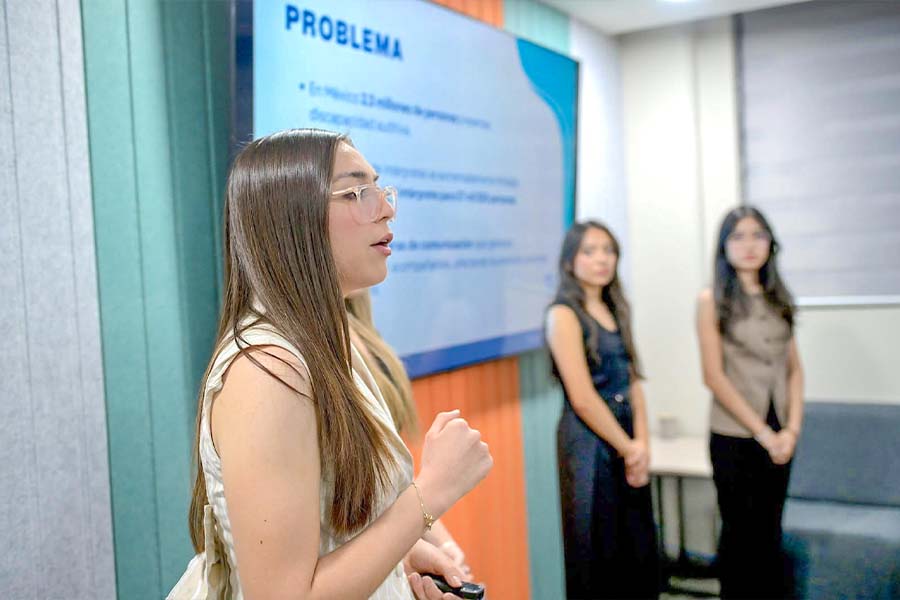 Mujer adolescente hablando, en el fondo se ve una pantalla con una presentaci&oacute;n y otras dos chicas a su derecha.  
