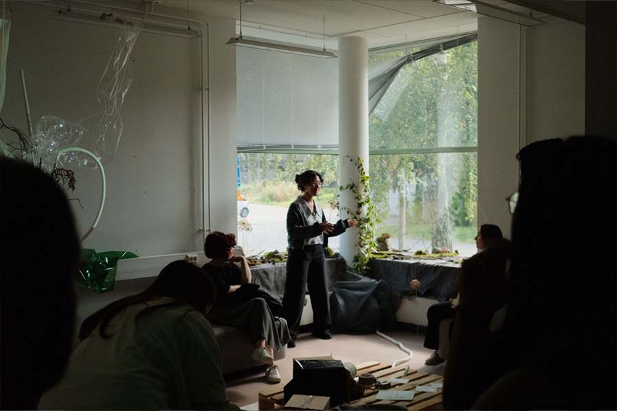 Estudiante en un cuarto oscuro, hablando de pie frente una ventana que deja entrar luz y la ilumina.