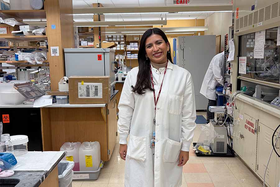 Joven vestida con bata de laboratorio, posando en el centro de un laboratorio de investigaci&oacute;n.