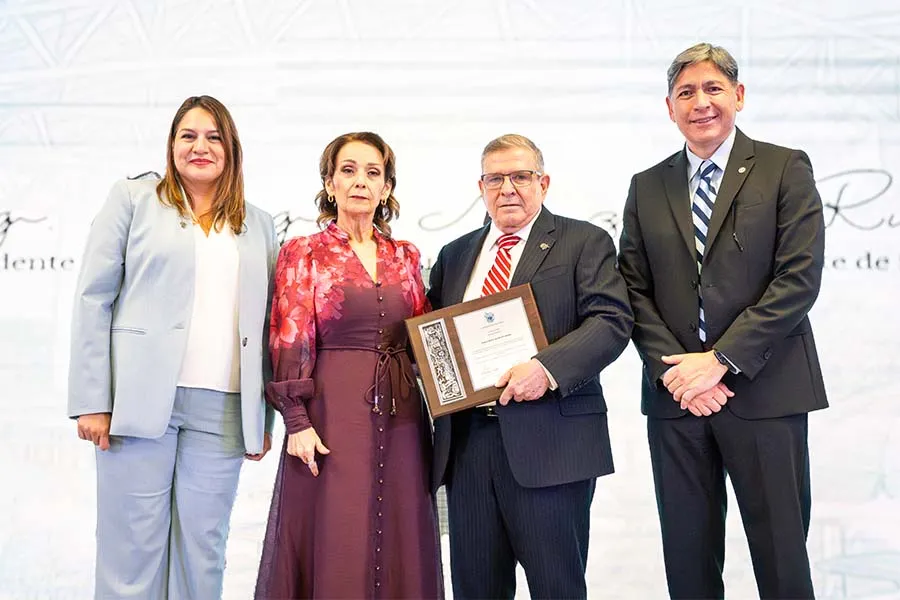 Dos mujeres y dos hombres posando frente a un fondo blanco. El hombre al centro de la foto sostiene un reconocimiento.