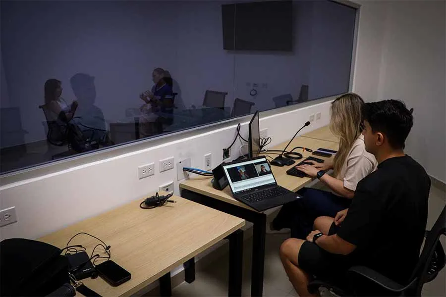 Dos estudiantes están del otro lado de una ventana a través de la cual se ve un grupo de personas sentadas en una mesa redonda de cristal.