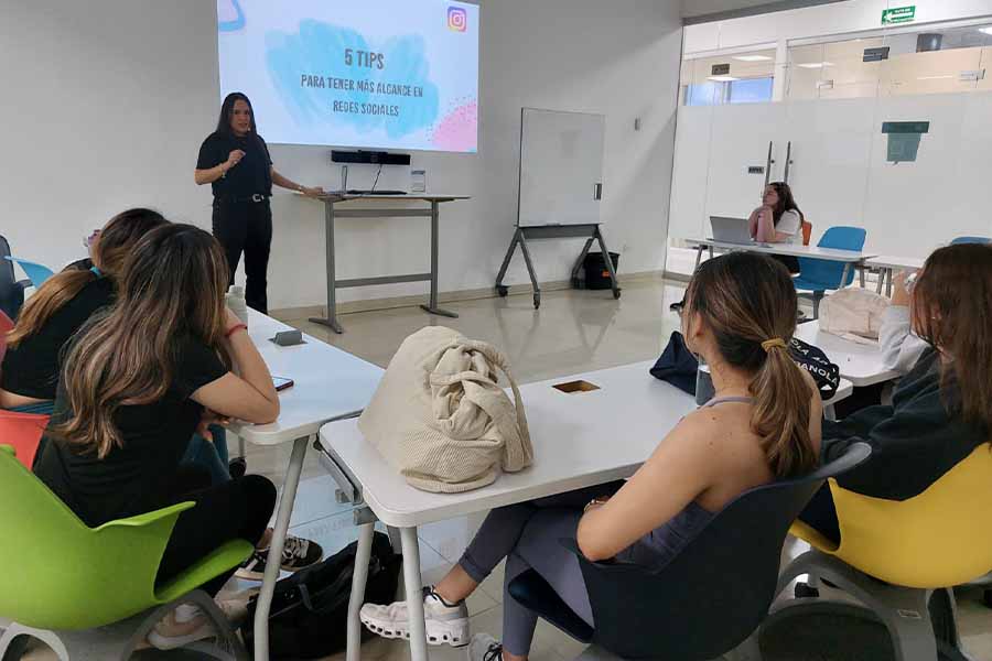 Mujer al frente de un salón de clases, hablándole a un grupo de alumnas. 