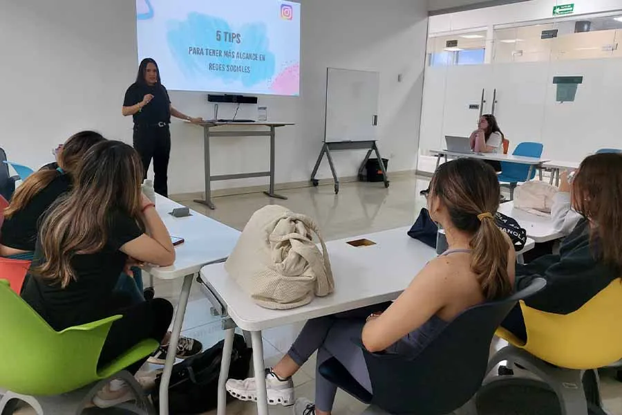 Mujer al frente de un salón de clases, hablándole a un grupo de alumnas. 