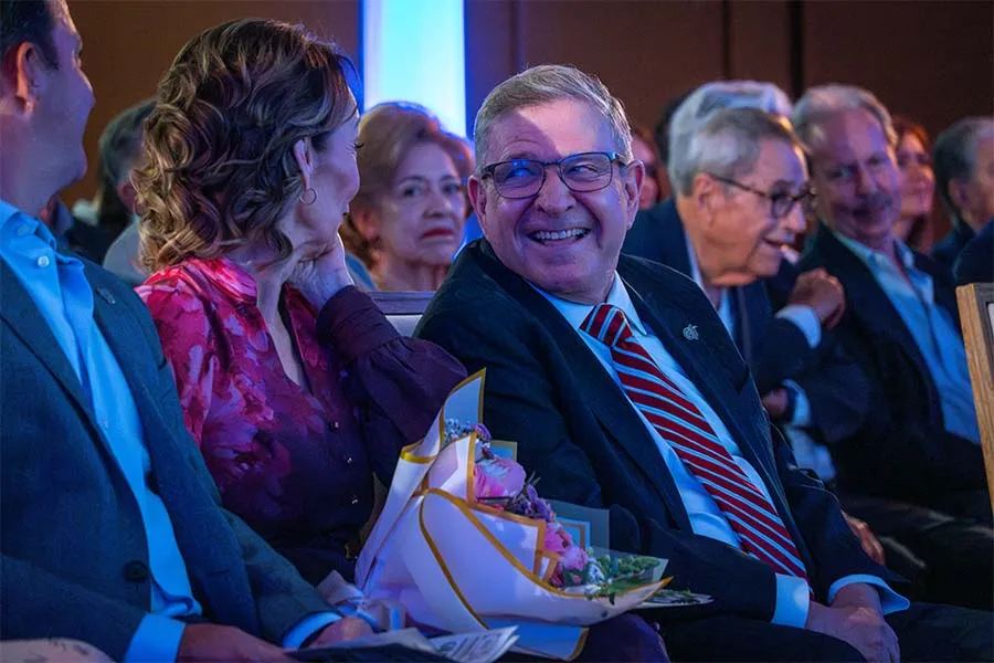 P&uacute;blico sentado en sillas. Al centro est&aacute; enfocando un hombre ri&eacute;ndose y volteando hacia la mujer que est&aacute; sentada a su lado. 