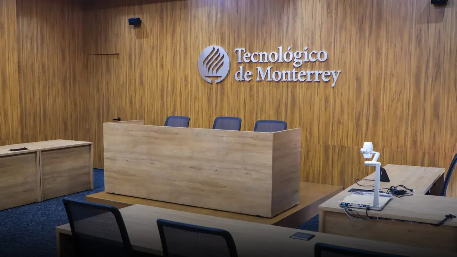 Vista de una sala de juicios con paredes recubiertas en madera, el escritorio del tribunal está al centro y en la pared detrás se ve el logo del Tecnológico de Monterrey.