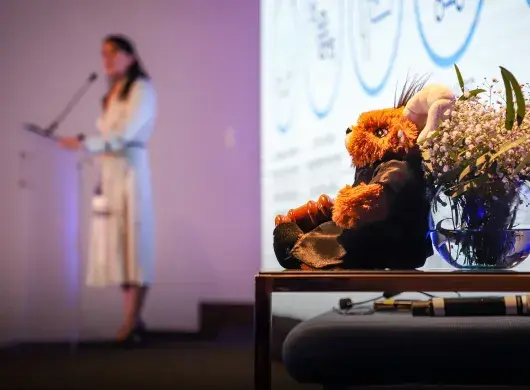En primer plano se ve un muñeco de un borrego vestido gon una bata negra y sosteniendo un mazo de madera, en el fondo se ve la figura borrosa de una mujer hablando en un escenario.