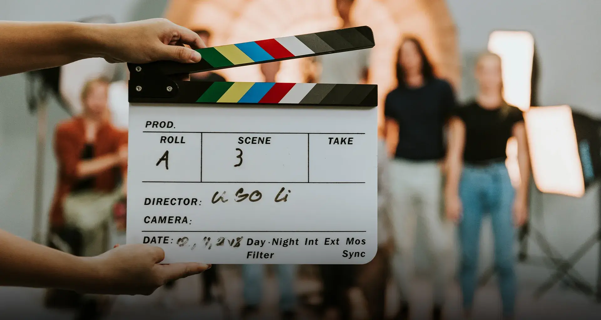 Primer plano de una claqueta de cine, al fondo se ve un grupo de personas desenfocadas.