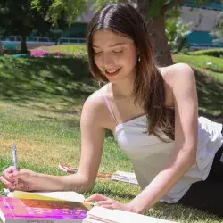 Alumna escribiendo al aire libre con libros a su alrededor