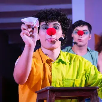 Actor con vestuario y nariz de payaso, sosteniendo un avión de papel.