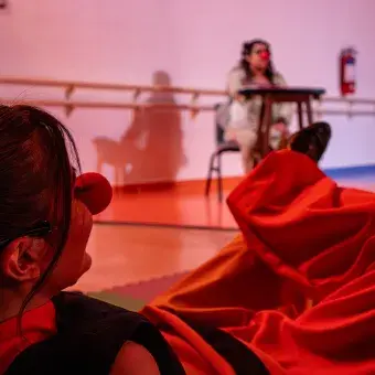 En primer plano se ve una chica acostada en el suelo, en el fondo se ve una actriz con vestuario y nariz de payaso en un triciclo.