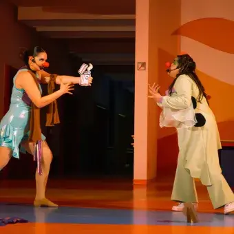 Dos actices con vestuarios y narices de payaso, una sostiene un dibujo de una botella atomizadora y lo apunta hacia la otra.