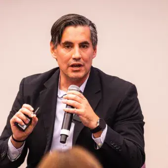 Luis Enrique Portales, durante su participación en la ceremonia NOVUS, expone datos y resultados del impacto de la innovación educativa mientras sostiene un micrófono y un control de presentación frente al público.